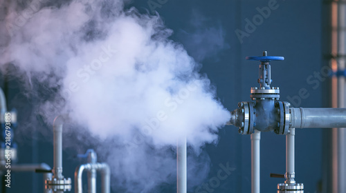Industrial safety scene showing pressurized gas escaping from metal valve and pipeline inside plant with visible vapor release