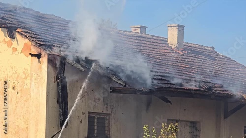 Firefighters extinguish burning house roof with water hose.