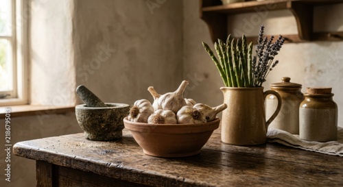 Rustic wooden table featuring a bowl of garlic bulbs, fresh asparagus, and lavender in a pitcher. Vintage farmhouse kitchen scene with mortar and pestle under natural window light.