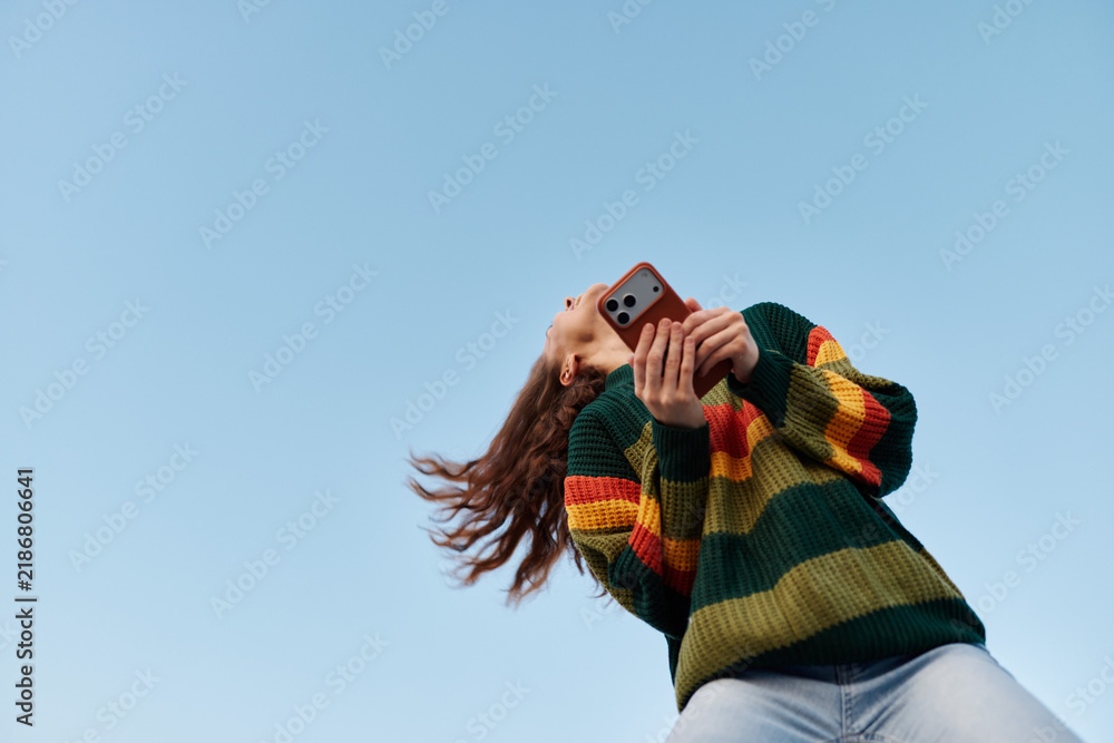 Obraz premium Low angle view of a young woman in a colorful striped sweater photographing with her smartphone against a clear blue sky. Concept of mobile photography and freedom