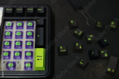 A top-down view of a mechanical numpad assembly displaying purple hot-swap switches. Black keycaps with distinct lime green legends and accents are scattered nearby, alongside a standar
