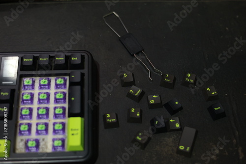 A top-down view of a mechanical numpad assembly displaying purple hot-swap switches. Black keycaps with distinct lime green legends and accents are scattered nearby, alongside a standar