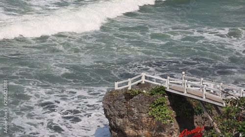 Rugged cliff meets crashing waves beneath a white railing. Salt spray drifts as surf pounds the rocky shore. Nature's power looms near human-made safety. Serene path invites pause by the roaring sea