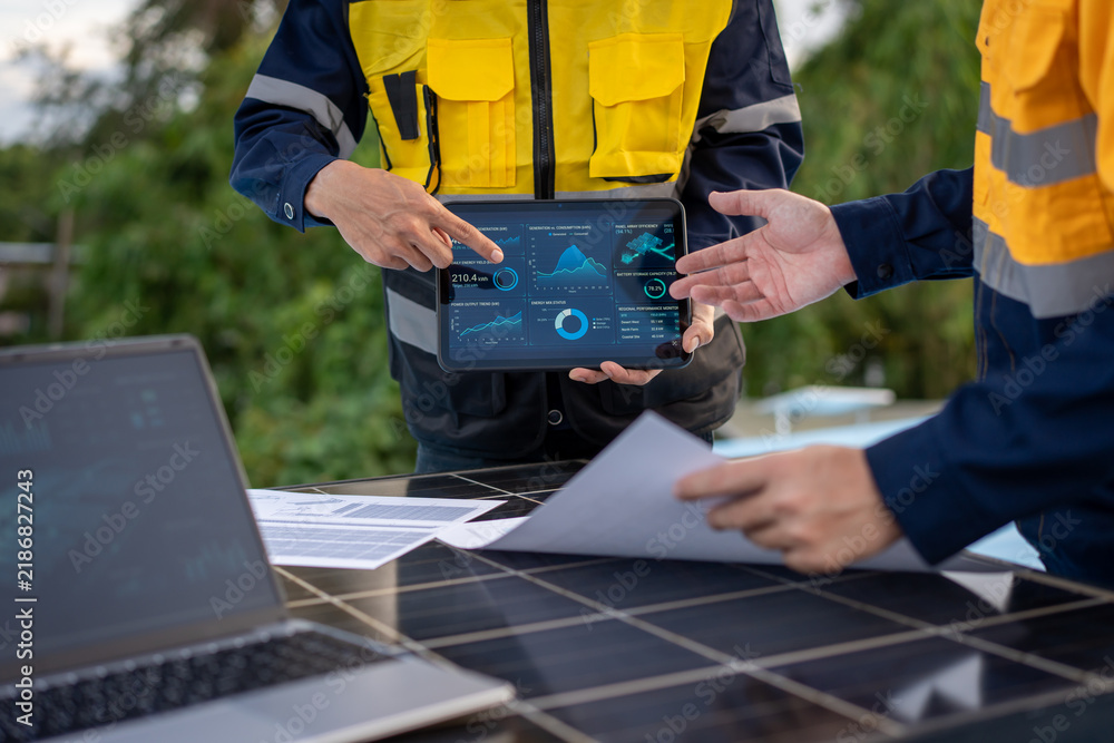 Obraz premium Solar energy technicians in safety uniforms review digital data on a tablet beside rooftop solar panels, with a laptop and paperwork during an outdoor inspection.