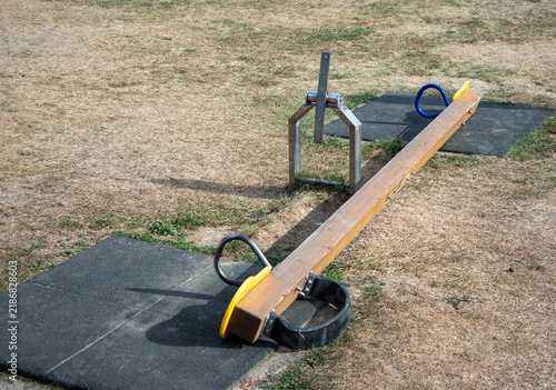 Broken and unusable wooden playground seesaw with a damaged central mechanism on dry grass, showing neglected play equipment, maintenance problems and a safety hazard.
