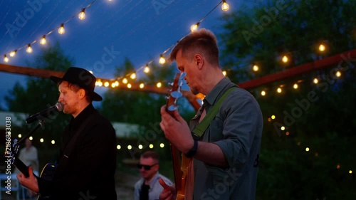 Live outdoor band performance under string lights at twilight with guitarist and singer playing music on stage during an intimate evening gathering