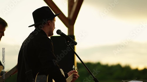 Outdoor country musician performing live acoustic guitar and vocals on riverside stage at golden hour sunset concert with backing band and scenic landscape