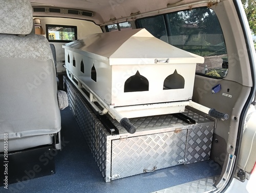 White Muslim funeral coffin or casket inside a funeral transport vehicle, featuring pointed-arch decorative openings inspired by Islamic architecture, a peaked lid, and a metal diamond-plate platform.