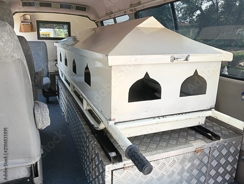 White Muslim funeral coffin or casket inside a funeral transport vehicle, featuring pointed-arch decorative openings inspired by Islamic architecture, a peaked lid, and a metal diamond-plate platform.