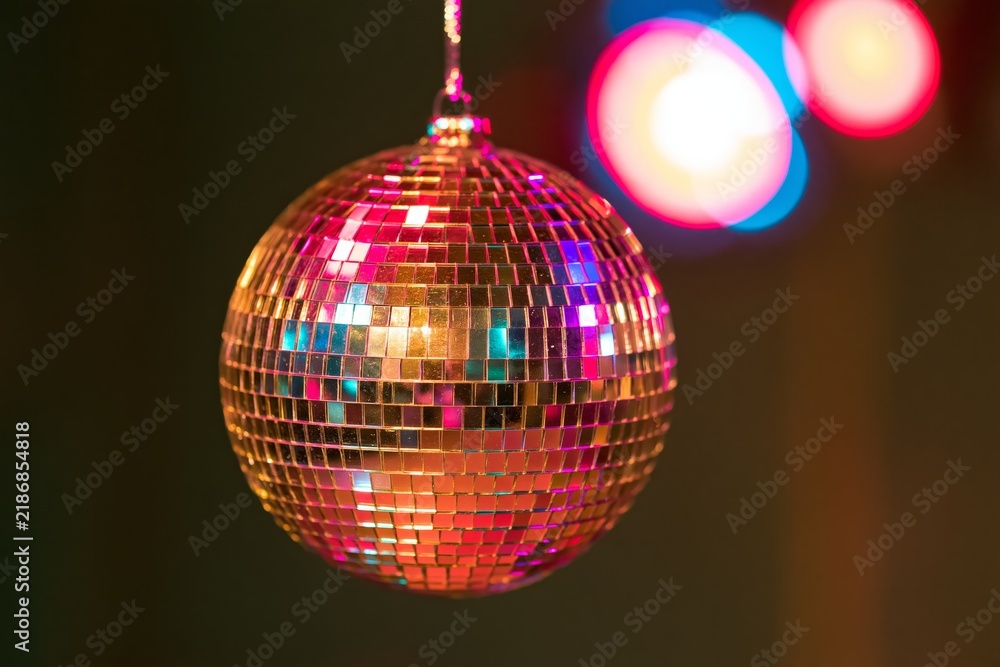Obraz premium Closeup of a faceted mirrored disco ball suspended against dark background with colorful bokeh lights reflecting magenta cyan and golden highlights across its surface creating vibrant festive