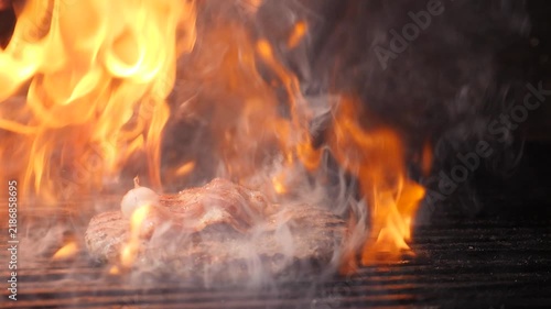 Juicy beef patty grilling on metal grate with intense orange flames and thick smoke rising. Dramatic dark background, professional restaurant kitchen cooking process. Slow motion 4k video
