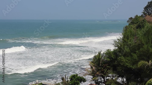 Ocean waves roll toward a lush, green cliffside. Tropical trees and palms frame the scene. Clear sky and calm waters suggest a peaceful day. Perfect for travel, vacation, or coastal lifestyle themes