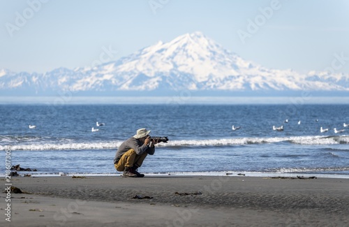 Nature photographer photographs birds on the beach, with white mountain peaks of Mount Redoubt in the background, mountains of the Aleutian range, Anchor Point, Anchor River State Recreation Area, Alaska, USA