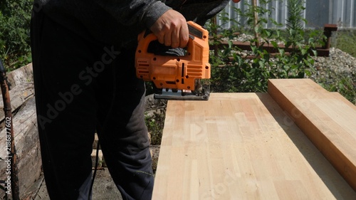 A man is cutting a wooden board with a bright orange electric jigsaw. The work is taking place outdoors, and fine sawdust is flying from the blade. A close-up shot focuses on the craftsman's hands