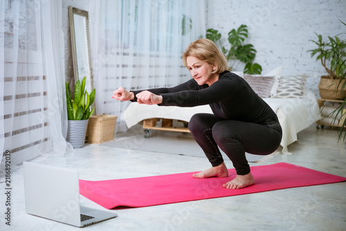 Canvastavla Mature female wearing black sportswear performing deep squat and balance exercise on pink yoga mat while watching instructional fitness video on laptop in bright room