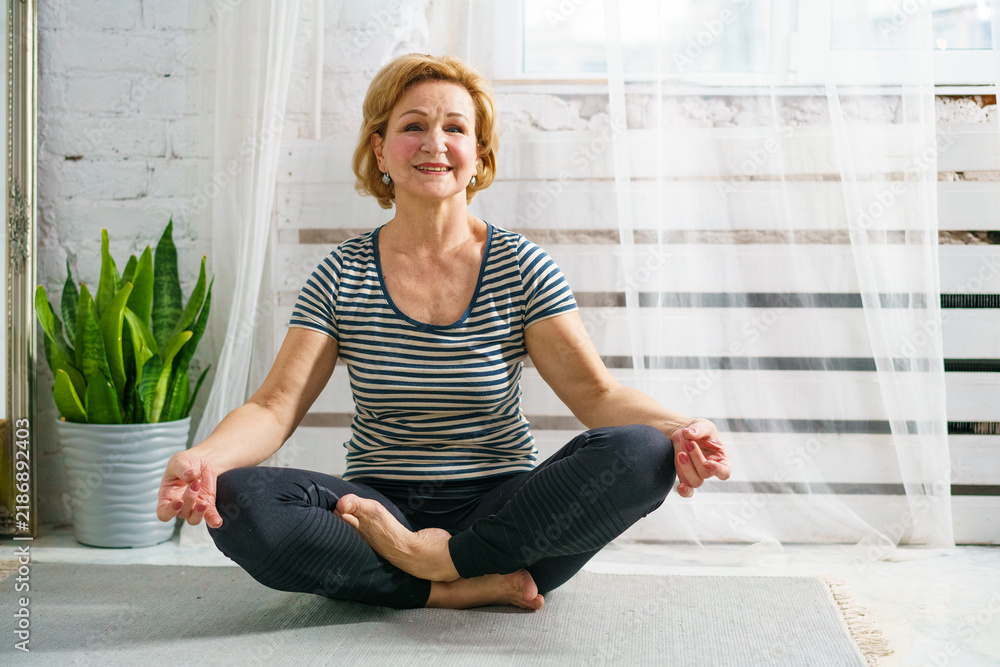Obraz premium Happy mature female in striped shirt sitting on floor mat in half lotus pose, smiling at camera. Positive healthy aging, morning vitality and joint mobility.