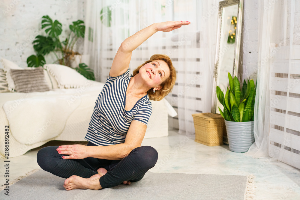 Fototapeta premium Happy mature female in striped shirt sitting cross legged on mat and doing lateral stretch. Cheerful senior woman practicing home workout and body flexibility.