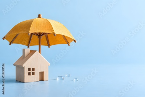 Holzhaus unter gelbem Regenschirm als Symbol für Versicherung und Schutz