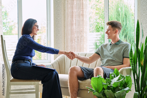 Female psychologist shaking hands with teenage boy sitting on sofa in office