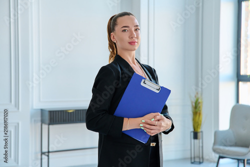 Confident businesswoman holding blue clipboard folder in modern office