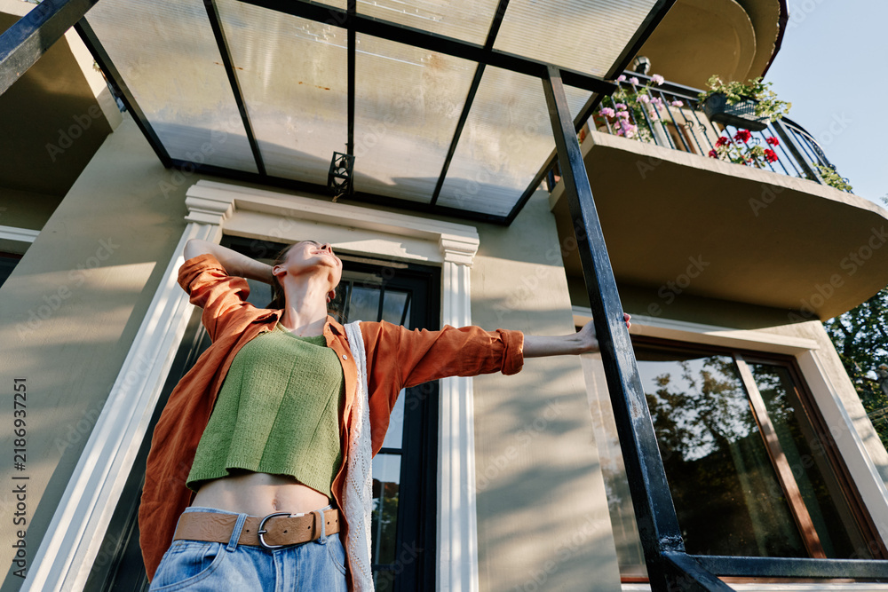 Obraz premium A joyful young woman stretches her arms wide outside a modern house with balcony and glass canopy, enjoying sunshine, freedom and happy summer lifestyle