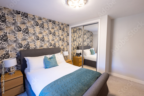 Contemporary double bedroom with grey upholstered bed teal bedding tropical leaf wallpaper mirrored sliding wardrobe wooden bedside cabinets lamps and soft neutral carpet