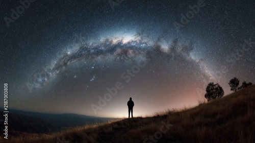 A solitary figure stands atop a hill, gazing at a breathtaking star-filled sky with the Milky Way arching gracefully above, surrounded by a serene natural landscape that evokes wonder and contemplatio