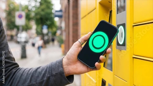Hand holding smartphone opening yellow parcel locker on city street. Contactless delivery pickup with green confirmation on phone screen.