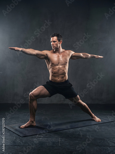 A powerful shot of a muscular man performing the Warrior II yoga pose on a mat in a dimly lit studio. This image captures dedication, balance, and the pursuit of a healthy, active lifestyle through...
