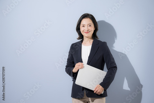 水色背景のスーツでパソコンを持つ笑顔の50代女性ビジネスイメージ、働く女性や転職やキャリアなどのカメラ目線笑顔