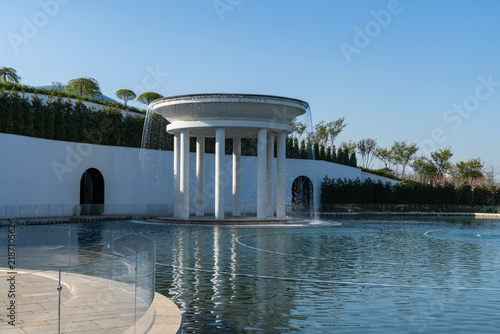 Circular rotunda fountain with water cascading down its sides into surrounding Lake of Love of calm water. Structure has white pillars supporting roof. Krasnodar. Galitsky Park - Cloud Park