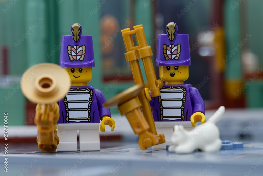 Fototapeta premium Tambov, Russian Federation - July 14, 2026 Two Lego marching band musician minifigures wearing purple uniforms and tall hats hold golden wind instruments and white cat on a city street.
