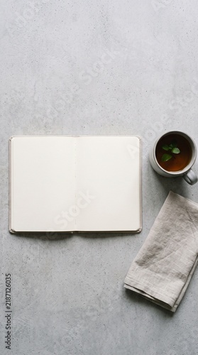 Open blank notebook, mug with tea and mint, and linen napkin on a grey concrete surface, top-down view.