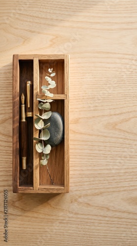 A minimalist wooden tray holding a fountain pen, eucalyptus sprig, and a smooth grey stone on a light wood surface.