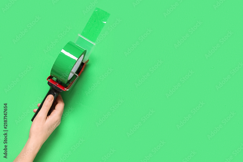 Obraz premium Logistics and shipping efficiency, hand holding a tape dispenser for packaging and parcel preparation on a solid green background