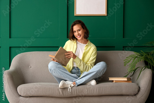 Pretty young woman reading book on sofa near green wall