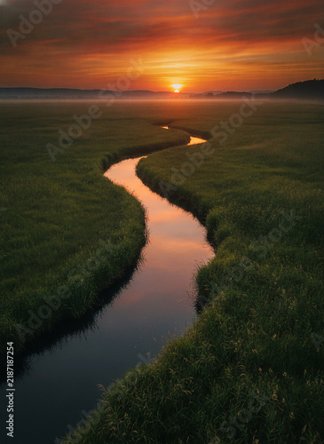 Aerial View Winding Stream Through Green Marshland at Golden Sunset, Misty Horizon, Reflective Water, Orange and Red Sky, Nature Landscape Photography