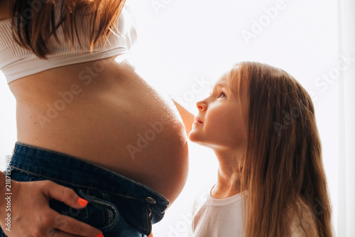Young daughter looking lovingly at her mother's pregnant belly in warm natural light. Tender family moment symbolizing motherhood, sibling love and anticipation of a new baby.