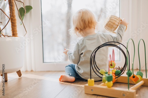 Little toddler sitting on the wooden floor by a large window and playing with a basket and colorful educational toys. Cozy candid childhood lifestyle scene with natural daylight at home.