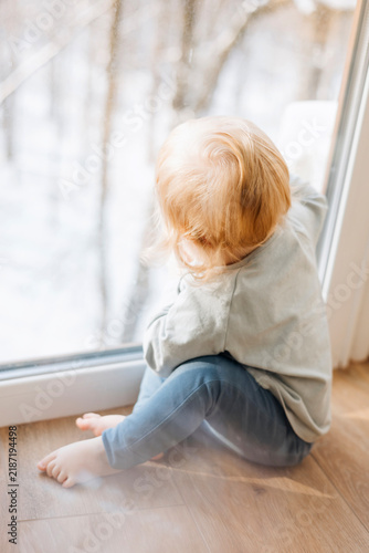 Rear view of a curious toddler sitting on a wooden floor by a large window and looking outside. Peaceful childhood moment at home with soft natural daylight, representing curiosity