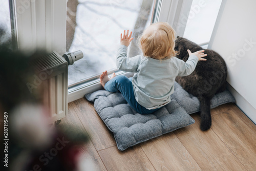 Little toddler sitting beside a cat on a cushion near a large window at home. Cozy candid moment showing friendship between a young child and a family pet in a bright modern interior.