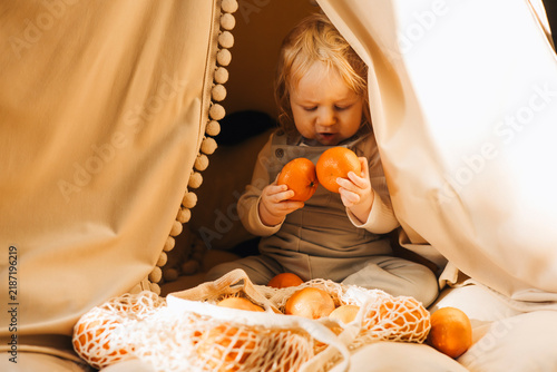 Cute toddler sitting inside a cozy blanket fort and exploring fresh oranges from a reusable mesh shopping bag at home. Candid childhood lifestyle scene with healthy food and imaginative play.