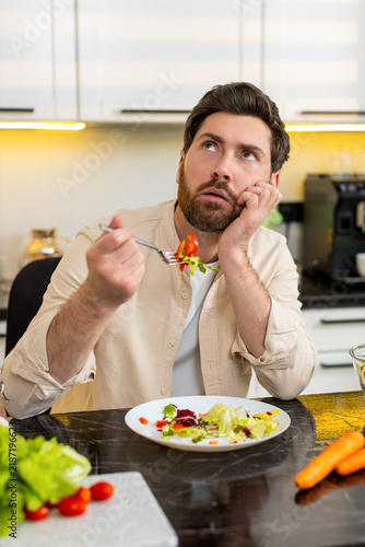 Canvas Print Middle-aged man tastes fresh vegetable salad in home kitchen feeling disappointed and forced to follow strict healthy diet plan