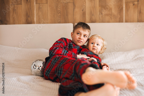 Two brothers wearing matching red plaid pajamas sitting together on a cozy sofa at home. Older boy gently embraces his toddler brother, showing friendship and family bonding during the holiday season