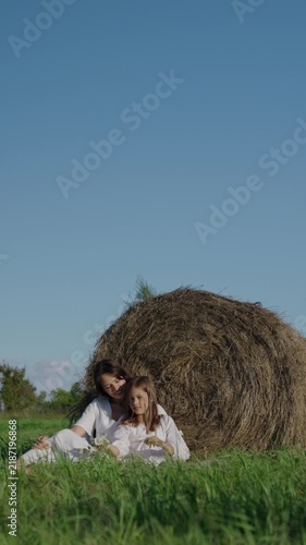A young mother and her 7 year old daughter, wearing white dresses, are sitting on a green meadow in a field next to a sheaf of hay, admiring the flowers on a sunny day. Vertical video.