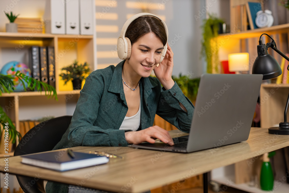 Fototapeta premium Young woman on home table works on laptop while listening to music in wireless headphones with focused calm mood. Adult girl on home table enjoys remote work and favorite songs during online duties.