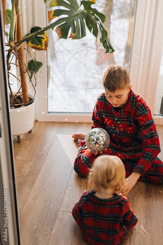 Two brothers in matching red plaid pajamas sitting by the window and playing with a silver Christmas decoration. Cozy sibling moment at home during the winter holiday season.