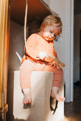 Curious baby sitting on a cat tree and playing with a hanging rope at home. Playful childhood moment in a cozy interior with warm natural sunlight