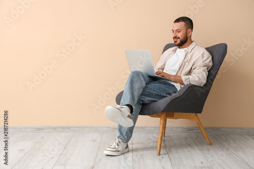 Young man sitting in armchair with laptop near beige wall in room