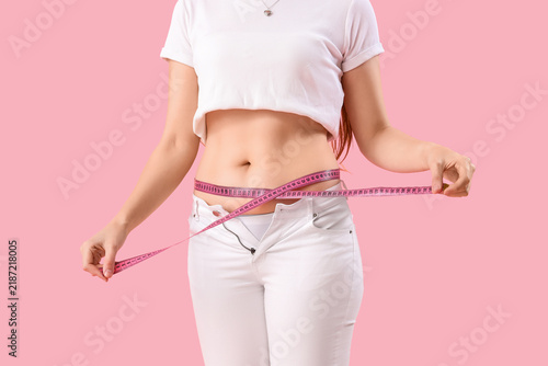Young woman in tight pants measuring her belly on pink background. Weight gain concept